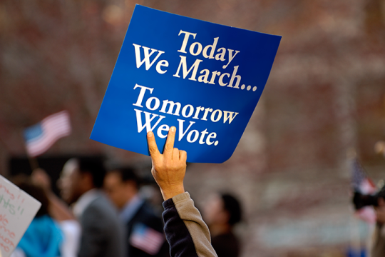 Photo of a hand holding up a blue sign with white writing that says "Today we March... Tomorrow we Vote"