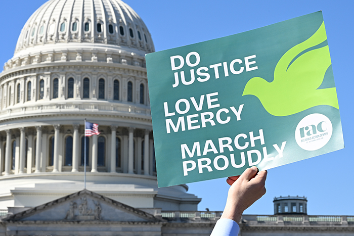Hand holding a sign that says Do Justice Love Mercy March Proudly, in front of the U.S. Capitol Building