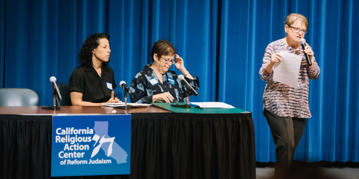 two women sitting behind a desk and one woman speaking with a microphone