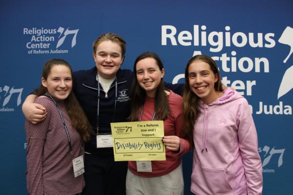 Four teens pose in front of a RAC banner holding a sign that says "I'm a Reform Jew and I support Disability Rights"