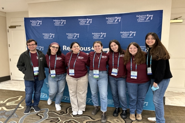 A group of seven Staff Assistants wearing matching maroon shirts and conference lanyards smiling and standing in front of a RAC banner.