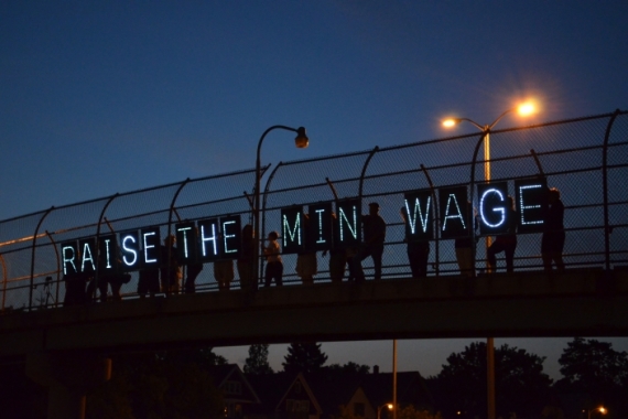 People standing on a bridge at dusk holding signs that spell out "Raise the Min Wage"
