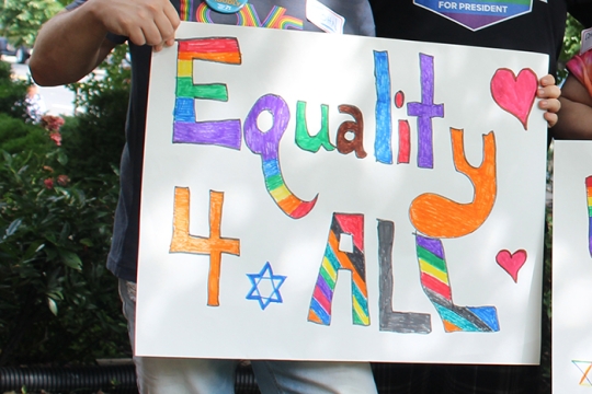 person holding a sign that says Equality 4 All in rainbow colors with a Star of David