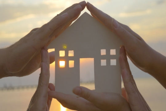 Cardboard cutout of house held up by hands; sunlight streaming in the windows