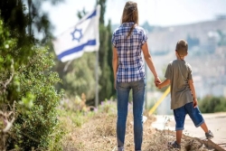 A woman holds a young childs hand with their backs toward the camera and an Israeli flag in the distance behind them