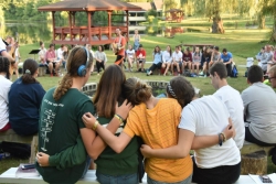 Campers sit on a bench with their arms around one another and their backs to the camera as they sit in a circle with camp in the background