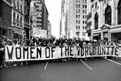 Black and white image of marchers holding a sign that reads WOMEN OF THE WORLD UNITE