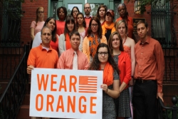 Group of somber people wearing orange clothing and holding a banner that says WEAR ORANGE